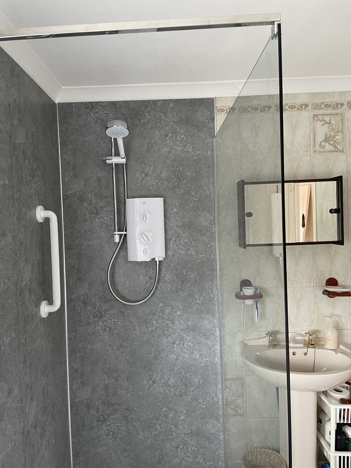 Shower area with an electric shower, grey wall panels, a grab rail and a glass screen