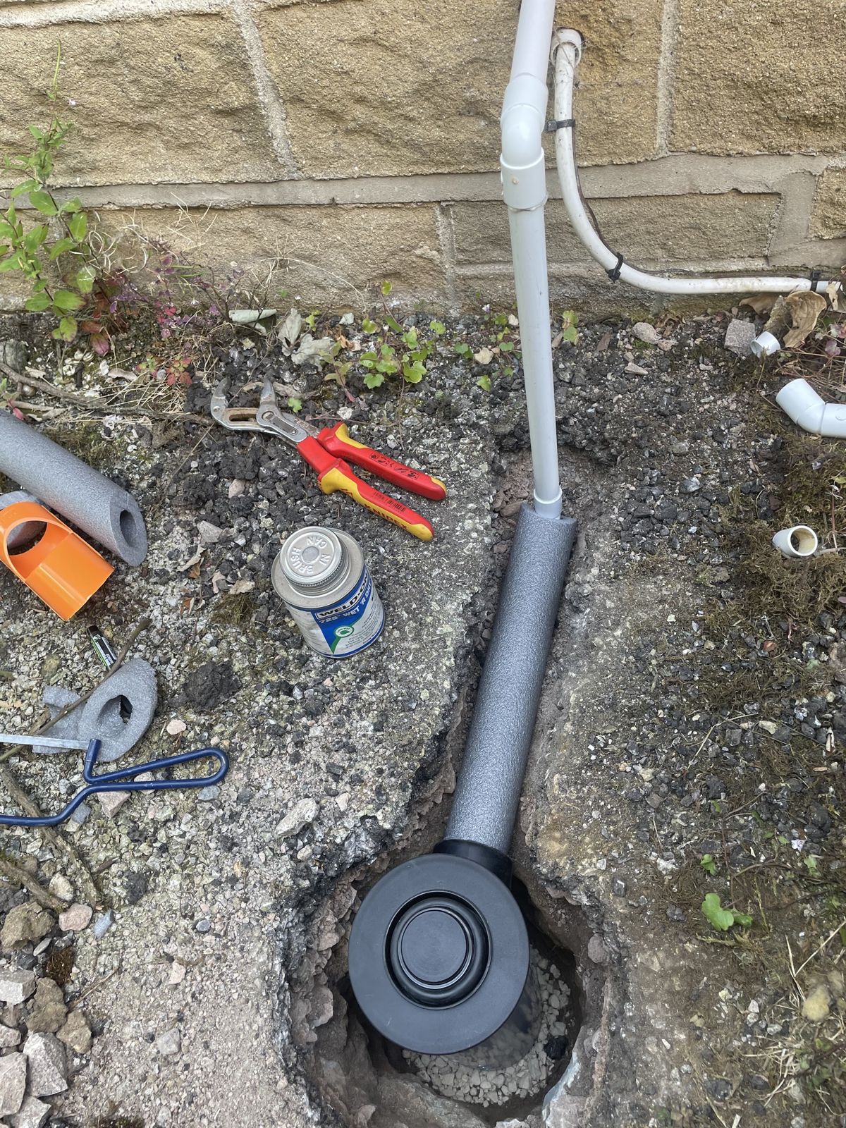 Insulated pipe laid in a dug trench beside a stone wall, with tools and offcuts